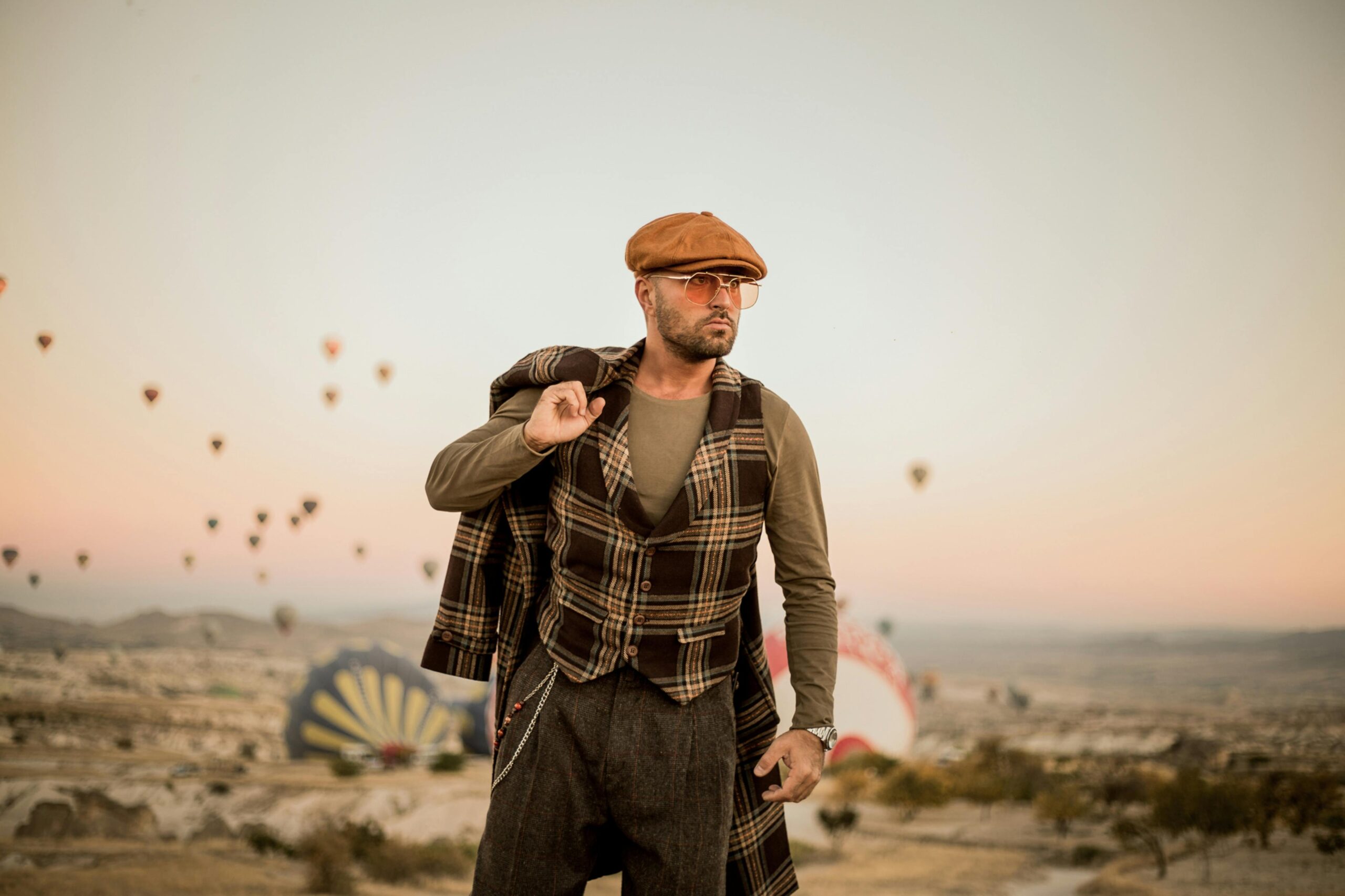 Stylish man wearing a plaid vest, flat cap, and tailored outfit standing outdoors with hot air balloons in the background